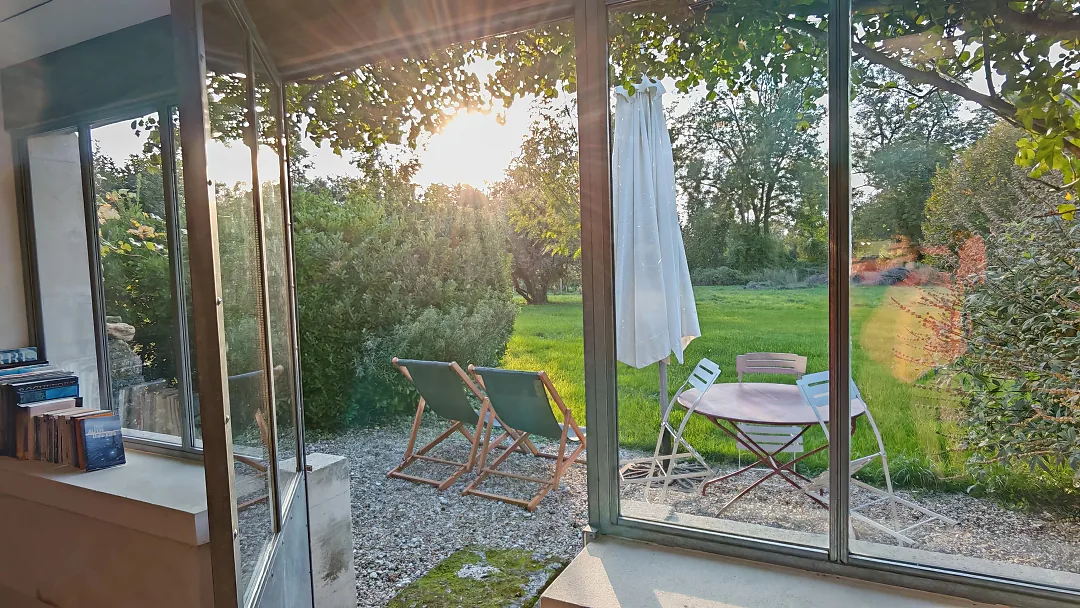 Le jardin de Borderies vu à travers la baie vitrée, transat et guéridon au soleil couchant